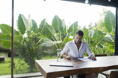 Mature man sitting at table in front of lush garden, writing - SBOF00908