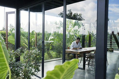 Mature man sitting at table in front of lush garden, writing - SBOF00909