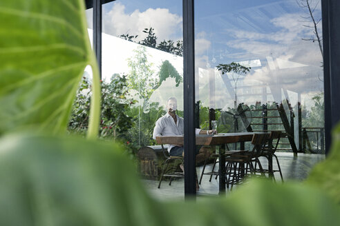 Mature man sitting at table in front of lush garden, relaxing - SBOF00911
