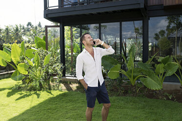Mature man walking in garden in front of modern villa, using smartphone - SBOF00914