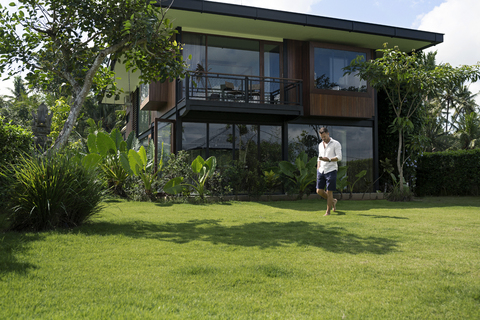 Älterer Mann, der im Garten vor einer modernen Villa spazieren geht und ein Smartphone benutzt, lizenzfreies Stockfoto