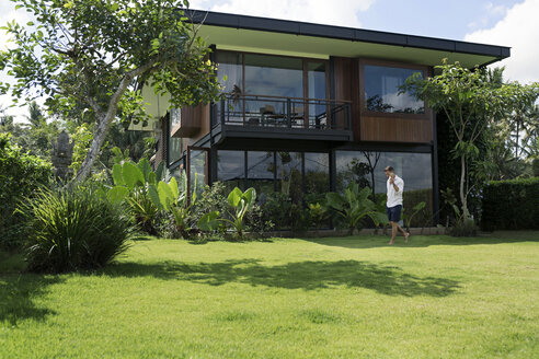 Mature man walking in garden in front of modern villa, using smartphone - SBOF00916