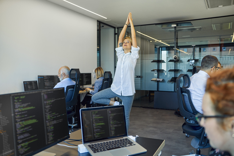 Frau übt Yoga im Büro, umgeben von beschäftigten Kollegen, lizenzfreies Stockfoto