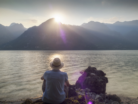 Italy, Lombardy, back view of man sitting at Lake Idro at sunset stock photo