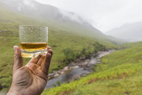 Great Britain, Scotland, Scottish Highlands, Glen Etive with River Etive and Glen Etive Falls, hand holding scotish whiskey in a glass - FOF09440