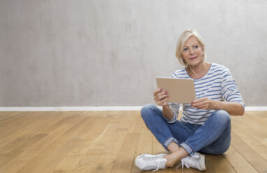 Portrait of smiling senior woman with tablet sitting on the floor - FMKF04620