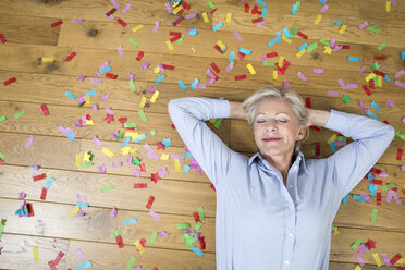 Portrait of smiling senior woman lying on floor covered with confetti - FMKF04627