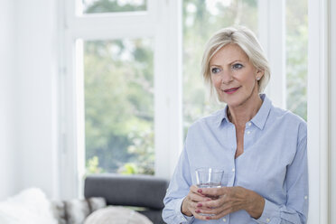 Portrait of smiling senior woman with glass of water in the living room - FMKF04635