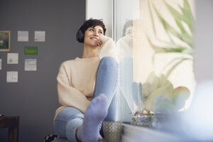 Lächelnde Frau zu Hause mit Kopfhörern am Fenster sitzend - RBF06104