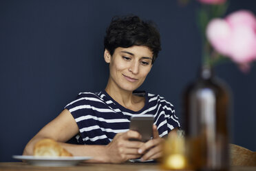 Lächelnde Frau zu Hause, die an einem Holztisch sitzt und ihr Mobiltelefon überprüft - RBF06145