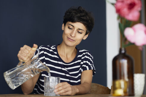 Woman at home sitting at wooden table pouring water into glass - RBF06146