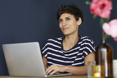 Porträt einer lächelnden Frau, die zu Hause an einem Holztisch sitzt und einen Laptop benutzt - RBF06147