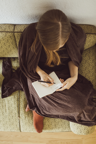 Frau sitzt auf der Couch im Wohnzimmer und schreibt in ein Notizbuch, Draufsicht, lizenzfreies Stockfoto