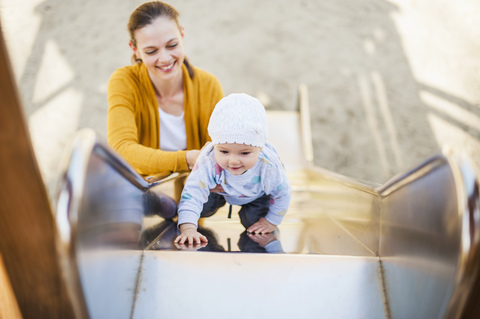 Kleines Mädchen klettert auf dem Spielplatz auf eine Rutsche und wird von ihrer Mutter gehalten, lizenzfreies Stockfoto