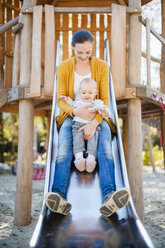 Happy baby girl sitting with her mother on shute on playground - DIGF03199
