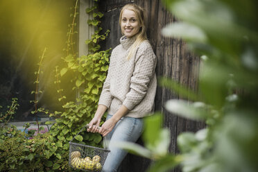Porträt einer lächelnden jungen Frau, die einen Drahtkorb mit Früchten im Garten hält - JOSF01887