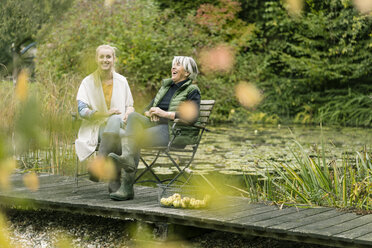 Glückliche junge Frau mit ihrer Großmutter auf dem Steg am Gartenteich sitzend - JOSF01891