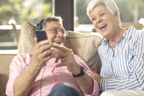 Glückliches älteres Paar mit Handy und Kopfhörern auf der Couch zu Hause - ZEF14728