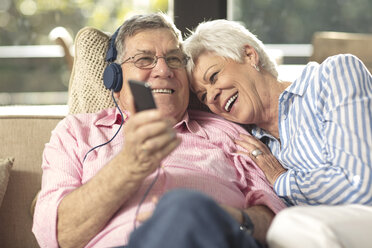 Glückliches älteres Paar mit Handy und Kopfhörern auf der Couch zu Hause - ZEF14729