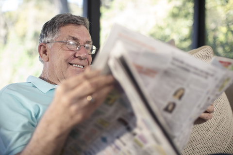 Lächelnder älterer Mann sitzt auf der Couch und liest Zeitung, lizenzfreies Stockfoto