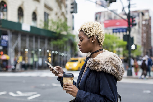 USA, New York City, modische junge Frau mit Kaffee zum Mitnehmen, die auf der Straße auf ihr Mobiltelefon schaut - MAUF01216