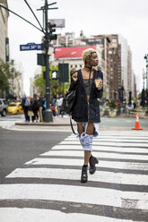 USA, New York City, modische junge Frau mit Kaffee zum Mitnehmen beim Überqueren der Straße - MAUF01218