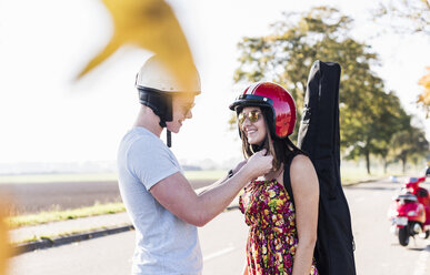 Young man closing helmet of girlfriend with motor scooter in background - UUF12256