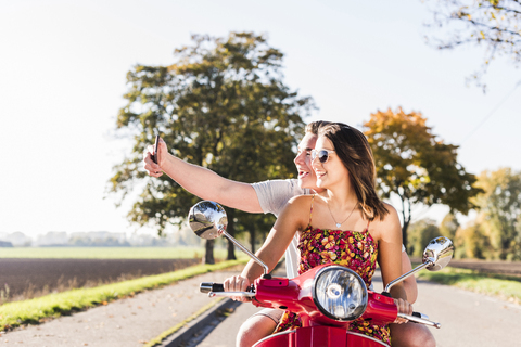 Glückliches junges Paar macht ein Selfie auf einem Motorroller auf einer Landstraße, lizenzfreies Stockfoto