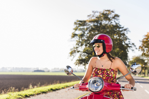 Glückliche junge Frau auf einem Motorroller auf einer Landstraße, lizenzfreies Stockfoto