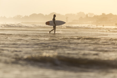 Indonesien, Bali, Surfer trägt sein Surfbrett bei Sonnenuntergang ins Meer - KNTF00918