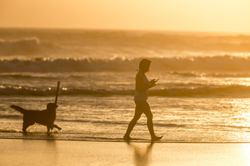 Indonesien, Bali, Silhouette einer Frau, die bei Sonnenuntergang mit ihrem Hund am Strand spazieren geht - KNTF00919