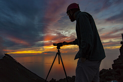 Italien, Äolische Inseln, Stromboli, junger Mann beobachtet Vulkanausbruch mit buntem Sonnenuntergang Hintergrund - THGF00026