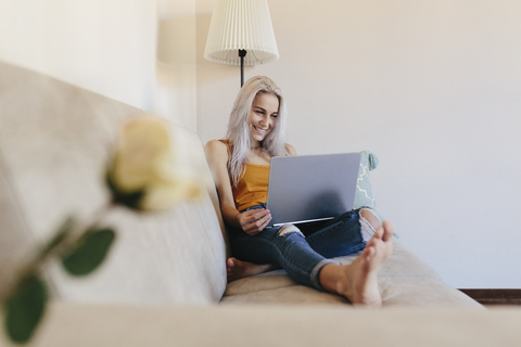 Lächelnde junge Frau mit Laptop auf der Couch zu Hause, lizenzfreies Stockfoto