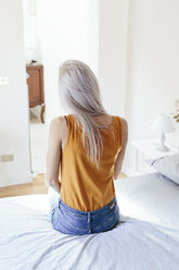 Rückansicht einer jungen Frau mit langen blonden Haaren, die zu Hause auf dem Bett sitzt - GIOF03366