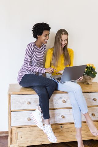 Zwei junge Frauen sitzen auf einem Schrank und schauen gemeinsam auf einen Laptop, lizenzfreies Stockfoto