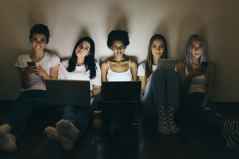 Eine Gruppe von Freundinnen sitzt zu Hause auf dem Boden und benutzt im Dunkeln Technik - GIOF03452