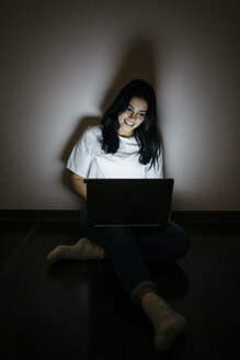 Junge Frau sitzt zu Hause auf dem Boden und benutzt einen Laptop in der Dunkelheit - GIOF03455
