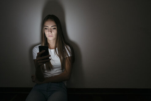 Junge Frau sitzt zu Hause auf dem Boden und benutzt ein Handy im Dunkeln - GIOF03457