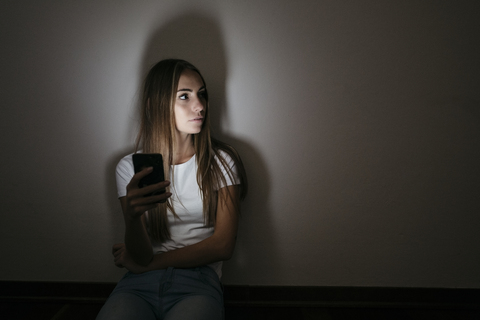 Young woman at home sitting on floor using cell phone in the dark stock photo