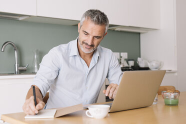 Lächelnder Mann, der in ein Notizbuch schreibt und einen Laptop im Heimbüro benutzt - ALBF00253