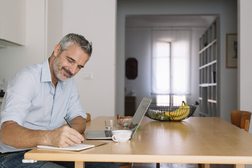 Lächelnder Mann, der in ein Notizbuch schreibt und einen Laptop im Heimbüro benutzt - ALBF00257