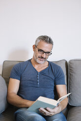 Mature man sitting on couch at home reading book - ALBF00271