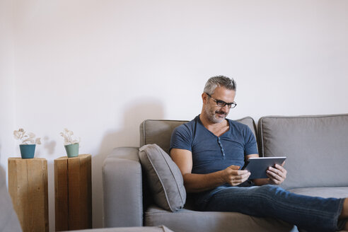 Mature man sitting on couch at home using tablet - ALBF00272