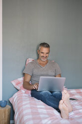 Lächelnder reifer Mann sitzt zu Hause auf dem Bett und benutzt einen Laptop - ALBF00276