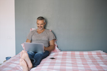 Lächelnder reifer Mann sitzt zu Hause auf dem Bett und benutzt einen Laptop - ALBF00277