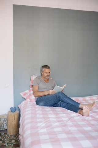 Älterer Mann sitzt zu Hause auf dem Bett und liest ein Buch, lizenzfreies Stockfoto