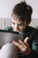 Portrait of laughing boy sitting on the couch in the living room using tablet - ALBF00305