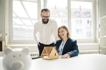 Lachende Frau und Mann mit Hausmodell und Sparschwein im Büro - JOSF01932