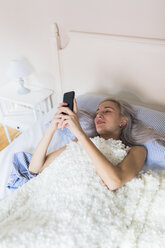 Smiling young woman lying in bed checking cell phone - GIOF03474