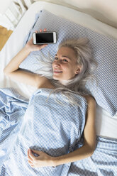 Lächelnde junge Frau im Bett liegend mit Handy in der Hand - GIOF03475